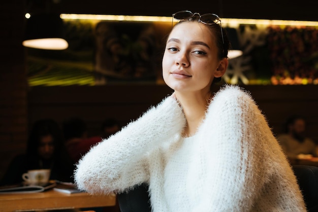 Hermosa joven independiente con suéter blanco descansando en el café del trabajo mirando a la cámara