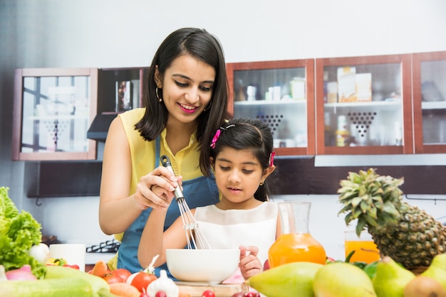 Hermosa joven india o asiática, madre e hija en la cocina, con mesa llena de frutas y verduras