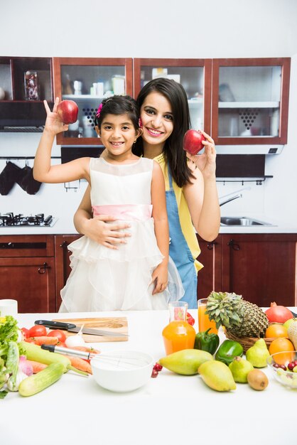 Hermosa joven india o asiática, madre e hija en la cocina, con mesa llena de frutas y verduras