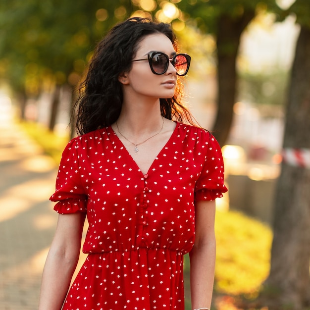 Hermosa joven italiana con estilo con el pelo rizado en ropa de verano de moda con vestido rojo vintage de moda y gafas de sol camina en la ciudad verde al atardecer