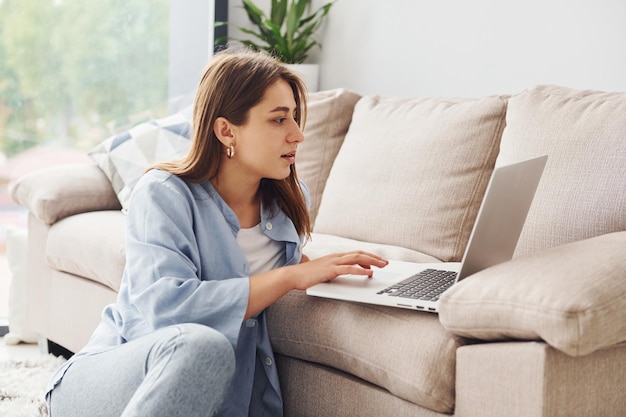 Hermosa joven en jeans y camisa azul acostada en el sofá con laptop