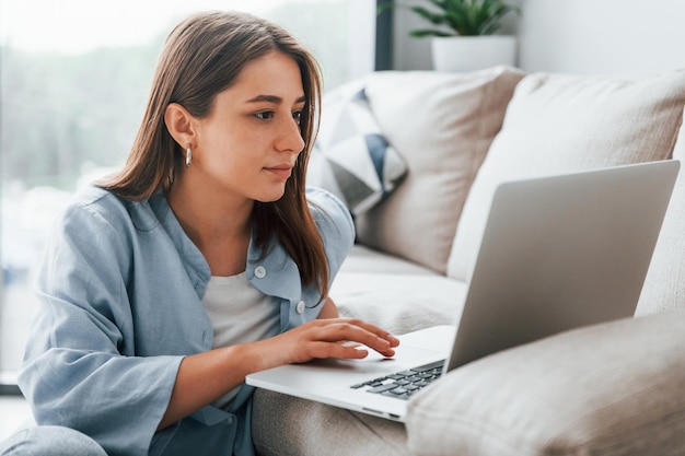 Hermosa joven en jeans y camisa azul acostada en el sofá con laptop