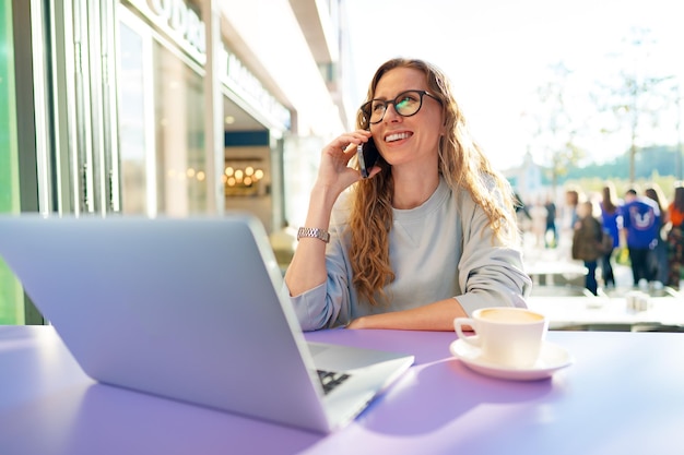 Hermosa joven con laptop hablando por teléfono en un café al aire libre