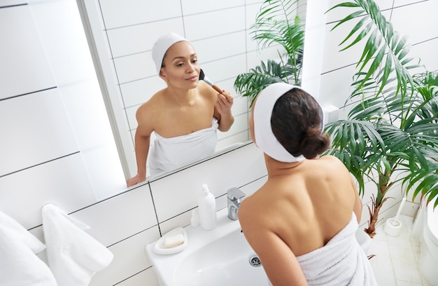 Hermosa joven latinoamericana aplicando su rubor o polvo facial ante el espejo.