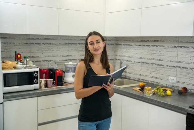 Hermosa joven lee un cuaderno en la cocina para cocinar alimentos saludables para el desayuno.