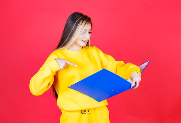 Hermosa joven leyendo documentos dentro de la carpeta azul en la pared roja