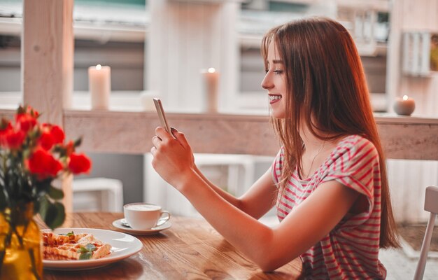 Hermosa joven leyendo un mensaje por teléfono mientras se relaja en un café y sonriendo