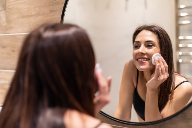 Hermosa joven limpiando su piel con un algodón, mirando al espejo en el baño de su casa.