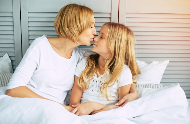 Hermosa joven linda madre está abrazando a su pequeña y encantadora hija. Mamá y niña acostadas en una cama blanca y relajándose juntas y divirtiéndose y pasando tiempo juntas