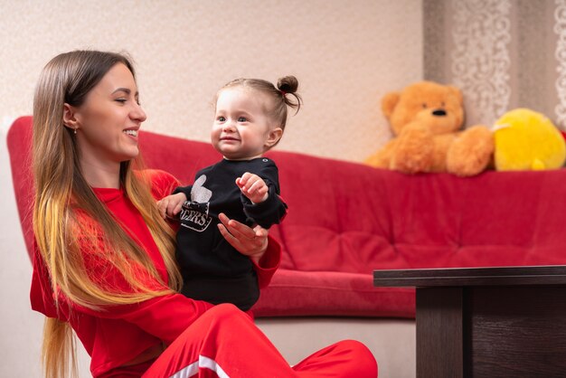 Hermosa joven madre con cabello largo y rubio sentada en un piso cerca del sofá rojo