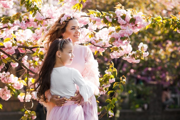Hermosa joven madre e hija miran hacia un lado cerca del árbol de sakura. Retrato familiar de primavera. Árbol floreciente.
