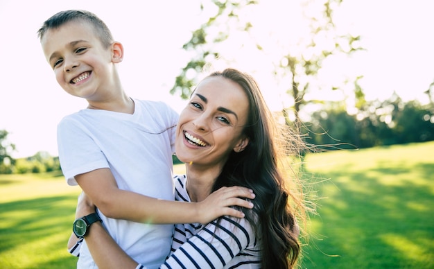 Hermosa joven madre encantadora con su pequeño y lindo hijo es divertirse y abrazarse mientras descansa sobre la hierba en el parque al aire libre