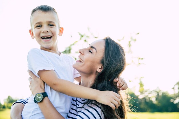 Hermosa joven madre encantadora con su pequeño y lindo hijo es divertirse y abrazarse mientras descansa sobre la hierba en el parque al aire libre