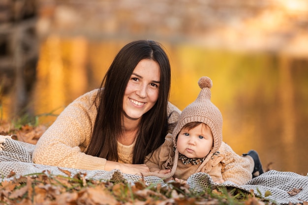 Hermosa joven madre jugando con su hija en una manta cerca del estanque en el parque de otoño en un día soleado