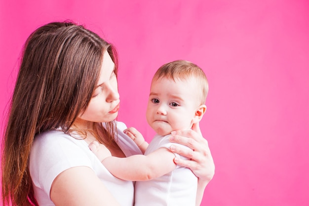 Hermosa joven madre mirando a su bebé, aislado en un fondo rosa. Madre joven con cabello largo castaño mirando a su hijo.