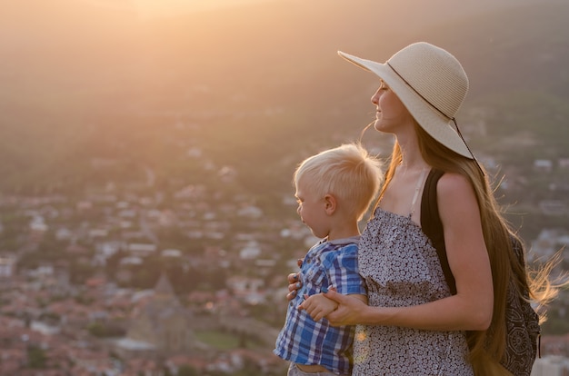 Foto hermosa joven madre con sombrero de paja y niño mirando la ciudad en puesta de sol. vista lateral