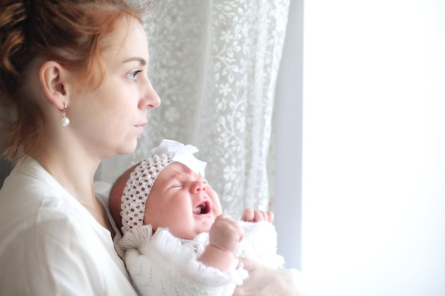 Hermosa joven madre y su bebé recién nacido en sus brazos de pie junto a la ventana