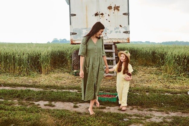 Hermosa joven madre con su hija en un vestido de lino con una canasta de refrigerios de fresas recoge una nueva cosecha y se divierte en el campo verde