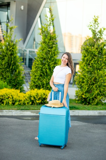 Una hermosa joven con una maleta azul frente al aeropuerto se va de vacaciones o viaja con un sombrero