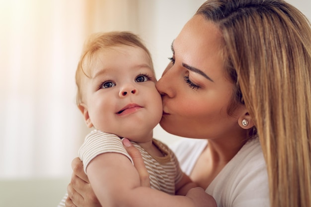 Hermosa joven mamá divirtiéndose con su lindo bebé en el hogar. Ella está besando al bebé.