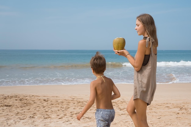 Hermosa joven mamá sosteniendo coco caminando en la playa con su hijo