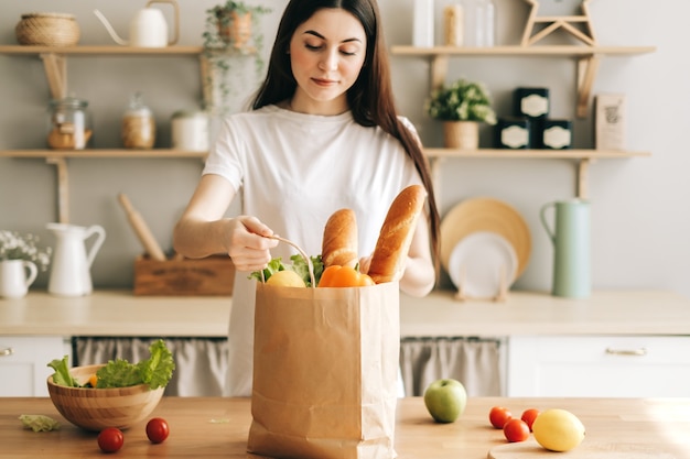 Hermosa joven mantenga eco bolsa de compras con verduras frescas y baguette en la cocina.