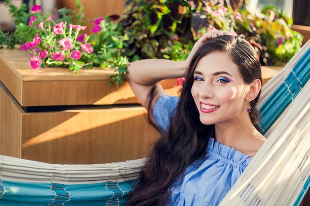 Una hermosa joven con maquillaje brillante y cabello largo y oscuro está sentada en una hamaca en la terraza de verano de un café de la ciudad mirando a la cámara y sonriendo