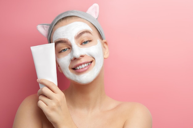 Hermosa joven con máscara de arcilla blanca en la cara y diadema coreana con orejas de gato sonríe y anuncia un producto cosmético aislado en un fondo rosa