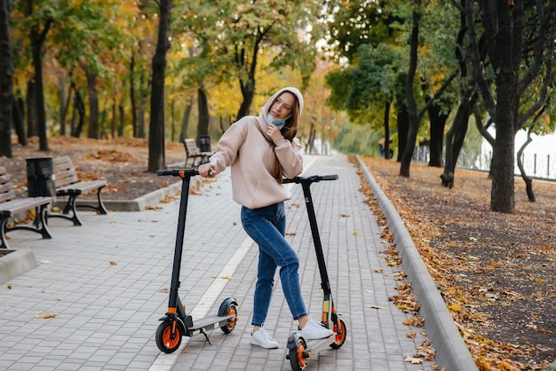 Una hermosa joven con una máscara está montando en el parque en un scooter eléctrico en un cálido día de otoño