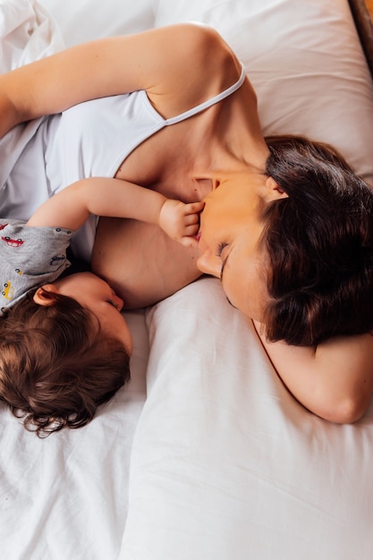 Hermosa joven de mediana edad amamantando a su hijo de un año acostado en la cama. lactancia. maternidad real