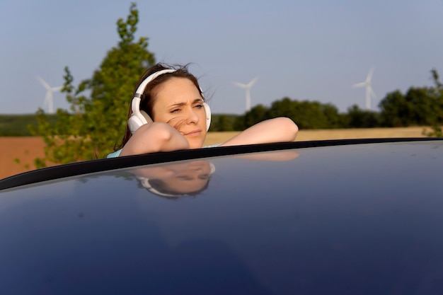 Hermosa joven de mediana edad escuchando música con auriculares apoyado en el techo del coche. molinos de viento en el fondo