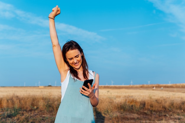 Hermosa joven de mediana edad mirando smartphone con molinos de viento en el fondo y levantando su brazo en señal de victoria