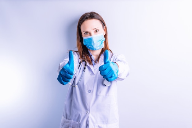 Hermosa joven médico con bata blanca y estetoscopio y máscara que muestra 2 dedos pulgadas al frente en modo de aprobación