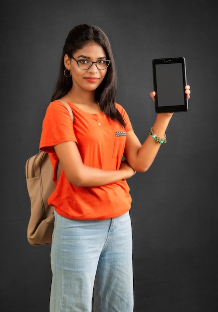 Una hermosa joven con una mochila muestra una pantalla en blanco de un teléfono inteligente o teléfono móvil o tableta posando sobre un fondo gris