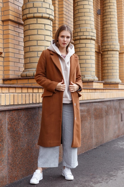 Hermosa joven en moda biege sudadera con capucha elegante abrigo de camello marrón y pantalones sobre fondo de pared de ladrillo caminando por la calle