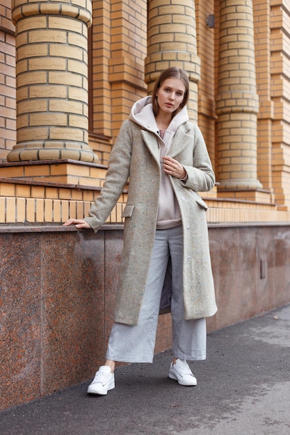 Hermosa joven en moda biege sudadera con capucha elegante abrigo gris y pantalones sobre fondo de pared de ladrillo caminando en la calle