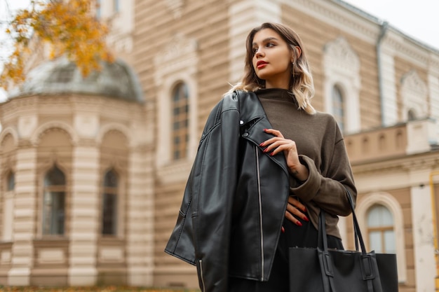 Hermosa joven de moda en una elegante chaqueta de cuero negro con un suéter verde con una bolsa camina en la ciudad de otoño con un edificio antiguo