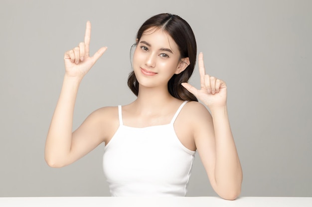 Hermosa joven modelo asiática apuntando al espacio de copia con piel limpia y fresca sobre fondo gris