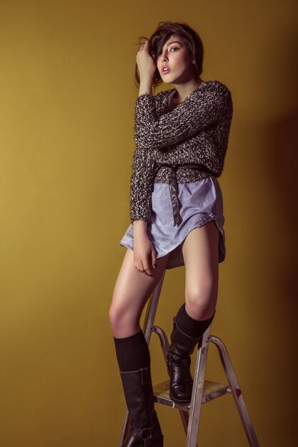 Hermosa joven modelo asiática en falda de mezclilla azul y blusa negra y maquillaje de moda posando sentado en la escalera y mirando a cámara. Tiro del estudio. Foto de alta costura.