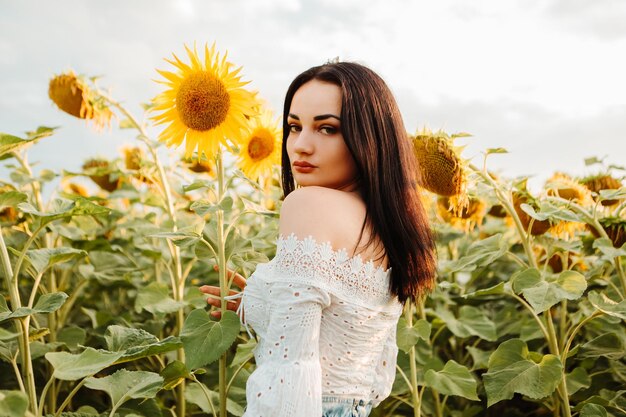 Hermosa joven modelo camina a través del campo de girasol floreciente ramo amarillo al aire libre puesta de sol