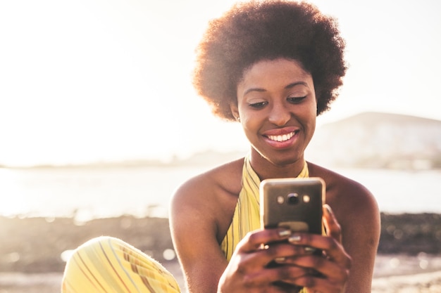 Hermosa joven modelo chica pelo negro rac africano utiliza tecnología de telefonía móvil para escribir amigos durante unas vacaciones. océano y luz de fondo, actividad de ocio al aire libre moderno