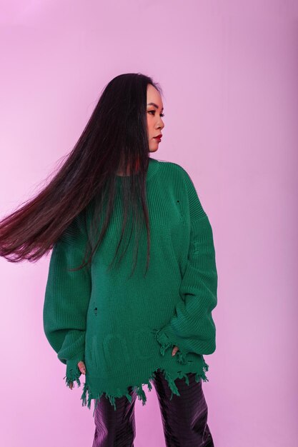 Hermosa joven modelo elegante con cabello saludable en un suéter de punto verde de moda con pantalones de cuero sobre un fondo verde en el estudio