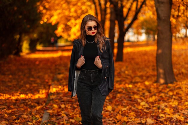 Hermosa joven modelo en un elegante traje con un suéter negro y una chaqueta camina en el parque con follaje otoñal