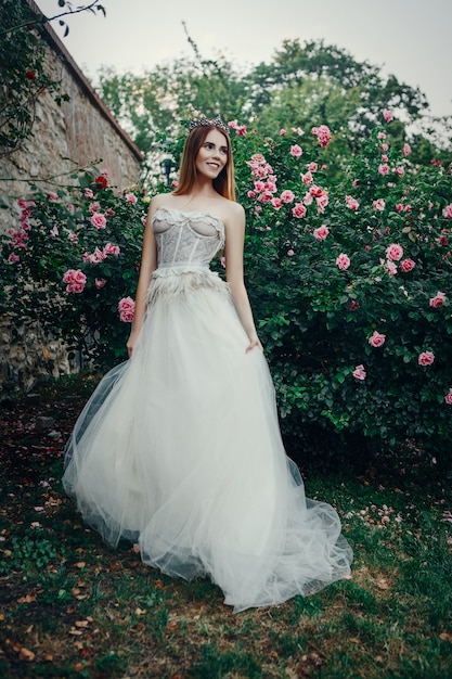 Hermosa joven modelo está posando con un vestido largo de marfil en el jardín con una corona en la cabeza