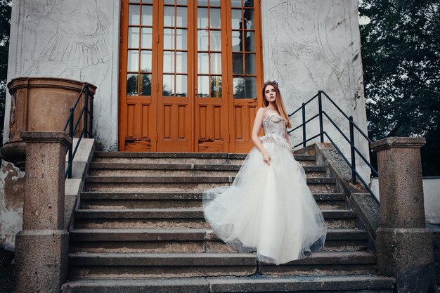 Hermosa joven modelo está posando con un vestido largo de marfil en el jardín con una corona en la cabeza