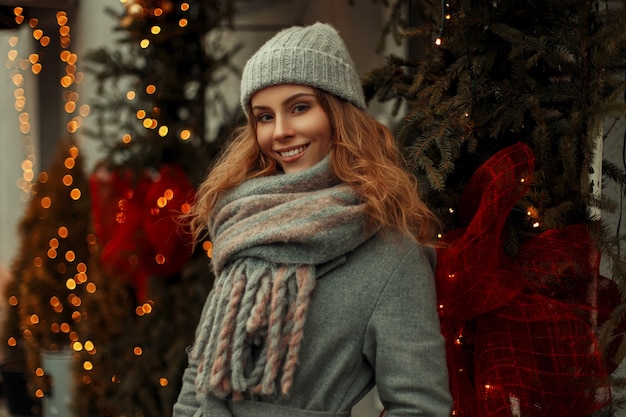 Hermosa joven modelo feliz con una sonrisa en un elegante abrigo gris y un sombrero vintage de punto de moda con una bufanda caminando en la ciudad de vacaciones cerca de las luces doradas