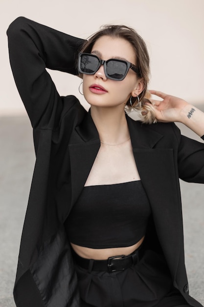 Hermosa joven modelo hipster con gafas de sol modernas y elegantes en ropa negra de moda elegante con top de chaqueta y pantalones sobre un fondo blanco en la calle