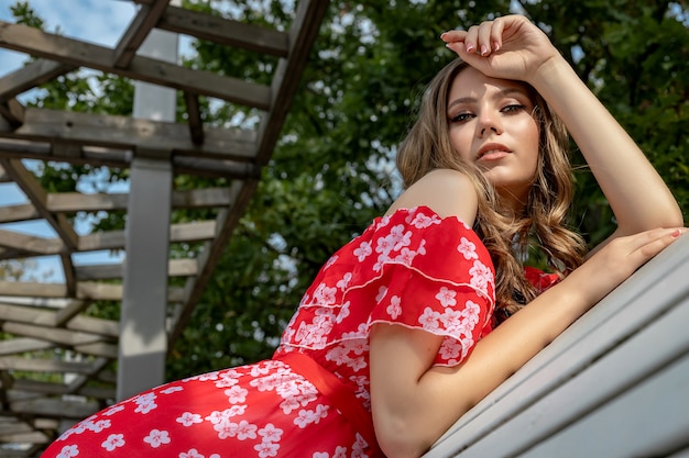 Foto hermosa joven, modelo, mujer de cabello castaño con cabello largo en un vestido rojo se sienta en un banco y posa con una mirada relajada y lánguida.