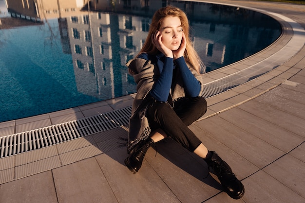 Hermosa joven modelo rubia se sienta junto a la piscina azul, disfruta del sol, los ojos están cerrados