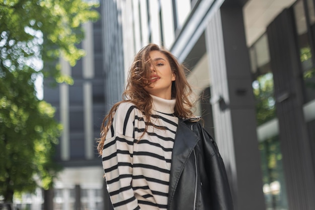 Hermosa joven modelo con un traje de moda con un suéter a rayas y una chaqueta de cuero camina en una ciudad moderna